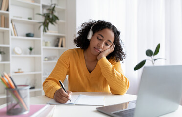 Obraz premium Bored young black female student having dull online lesson on laptop computer, writing down information at home