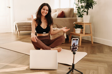 Athletic young millennial woman is engaged in sports training and exercise online in a room at home. A fitness blogger records videos during a workout using a smartphone and laptop. Selective focus