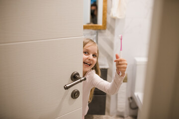 A smiling girl kid staying behind bathroom door with toothbrush in her hand.