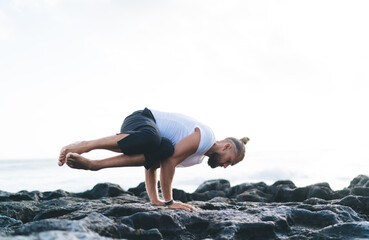 Strong Caucasian man standing on hands practice balance asana during morning time at rocky seashore, young male in sportswear have holistic therapy for recreating during active hatha training