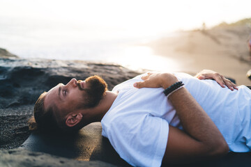 Bearded man with closed eyes lysing and resting during pastime for pranayama breathing in nature environment, calm male yogi feeling mindfulness enlightenment during holistic healing and retreat - Powered by Adobe