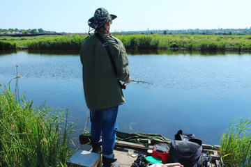 A fisherman with a fishing rod stands on a pier on the banks of the river, rear view on a sunny summer day among the reeds