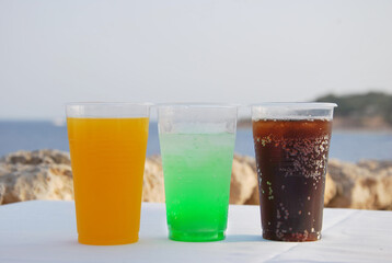 set of three plastic glasses refreshing carbonated cocktails standing on  table on  beach against blurred turquoise sea.