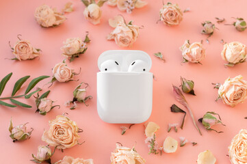 White wireless headphones in the case on playful pink background with delicate dry roses flowers.