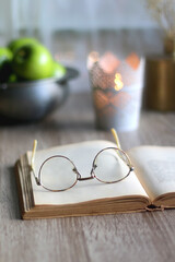 Bowl of green apples, open book, reading glasses, lit candle and flowers on the table. Selective focus.