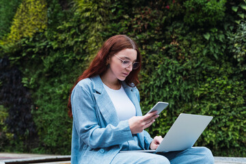 Woman reading text business message from her managers connected to public internet outdoors, beautiful professional woman chatting on cell smartphone and using laptop while relaxing outdoors