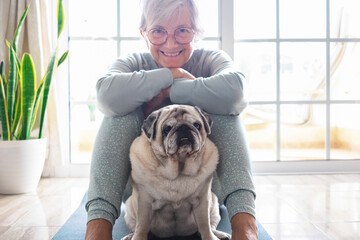 Close up portrait of funny old  pug dog looking at camera. Senior smiling fun woman sitting on floor ready to practice yoga exercises enjoying relax at home. Recreation with dog concept