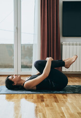 Beautiful Woman Practicing Yoga, Doing Apanasana Exercise at Home. 
Side view of woman in black sportswear relaxing in knees to chest pose on a exercise mat in a living room.