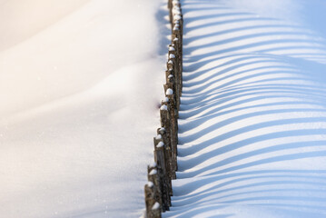 Old wooden fence covered with snow. Shadows from the sun on the snow.