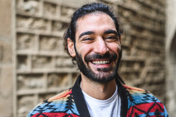 Merry Hispanic man in colorful jacket with beard and ponytail smiling and looking at camera. while...
