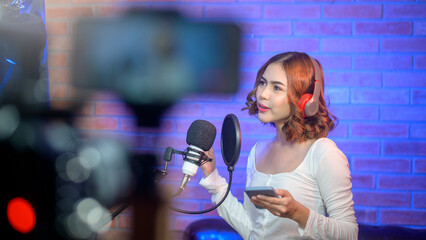 Young smiling female singer wearing headphones with a microphone while recording song in a music studio with colorful lights.