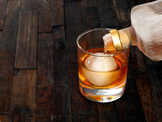 Liquor being poured over an artisan ice ball on a rustic wooden background. - obrazy, fototapety, plakaty