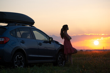 Silhouette of female driver standing near her car on grassy field enjoying view of bright sunset. Young woman relaxing during road trip beside SUV vehicle