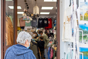 An unidentifiable senior woman with a mask walks into a souvenir store in Zahara de la Sierra, Spain as a masked family shops inside.