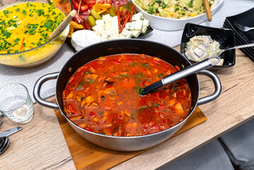 Goulash soup with pork and various vegetables, lying in a pot with a spoon on a table filled with food.