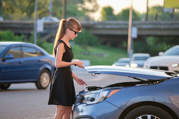 Young woman driver opening car bonnet inspecting broken motor on a city street. Vehicle malfunction concept