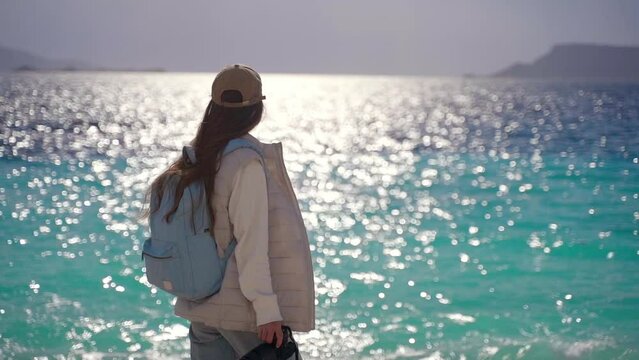 A woman with a backpack and a camera walks along the sea on the beach. Beautiful azure sea on the background