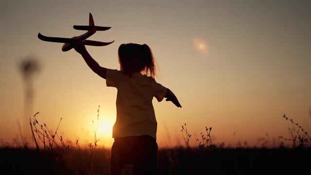 Happy girl with toy airplane in her hands. Silhouette of child in park in grass. Girl run across field with an airplane. Child pilot dream of flying. An active lifestyle in nature. Sunset sky in park