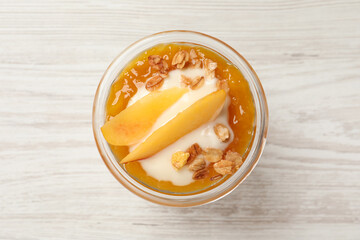 Tasty peach yogurt with granola, jam and pieces of fruit in glass on white wooden table, top view
