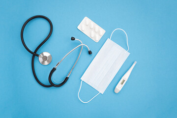 Top view, medical accessories on blue background. Stethoscope, medical mask, pills, medicines, thermometer. Flat lay.
