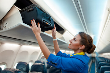 Woman flight attendant or air hostess placing travel bag in overhead baggage locker while standing...
