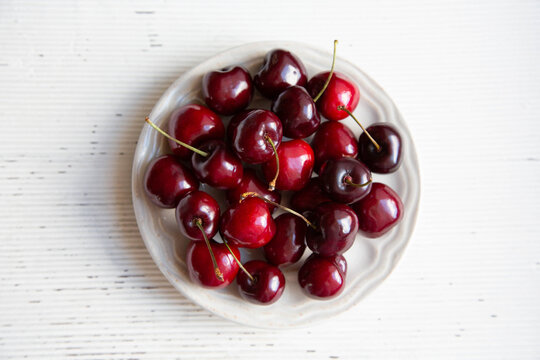 A ripe cherry on a white plate is on a wooden tray on a light background. The image is close-up and from the top point.