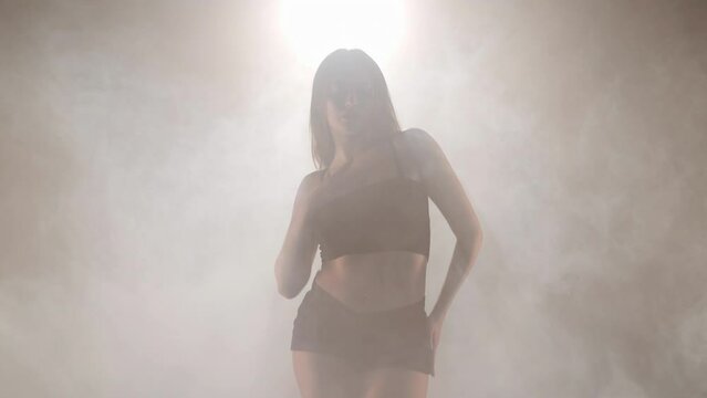 Artistic young woman dancing backlit fog smoke indoors. Coming out of smoke. Confident graceful Caucasian female dancer rehearsing modern performance with ballet movements. Choreography. Handheld