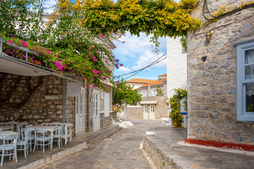 Fototapeta na wymiar A picturesque street of stone and whitewashed homes, pink flowers, and a sidewalk cafe in the picturesque village of the small Greek island of Hydra, Greece. 