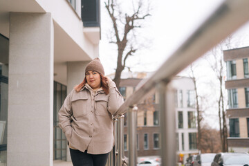 Portrait of confident overweight woman in warm hat and jacket standing near railing of building at urban street in cloudy autumn day, looking at camera. Freezing female waiting for someone outdoors.