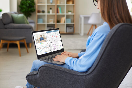 Young woman sitting in comfortable armchair at home, using modern laptop computer, organizing schedule in calendar planner, having conference call with business colleague, communicating in work chat