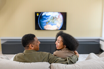 Back view of the caucasian couple in love sitting at the sofa and watching TV with pleasure smiles. Multiracial man embracing his lovely wife and looking at each other. Home concept