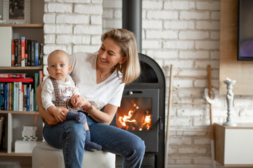 Portrait of smiling blond woman with baby in hands basking near comfort hot warm fireplace with firewood burning inside.