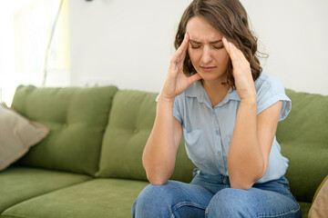 Obraz premium Upset young woman has strong headache, sitting on sofa at home and massaging temples, feels tired and exhausted. Sad lady worrying, feels anxiety about problems, suffers from migraine