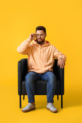 Handsome bearded man with eyeglasses sitting in leather armchair on yellow background