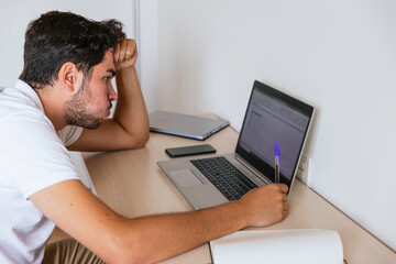 Side view of a young man puffing through his mouth, worried, sitting at a desk with his hand on his head with a computer taking notes. Concept: teleworking or having online classes