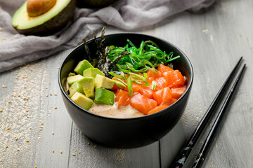 Poke with salmon, avocado and chukka with sauce and rice on black bowl on grey wooden table