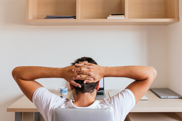Young man on his back with his hands clasped above his head looking at the computer, with a coffee. Concept: teleworking or having online classes.