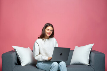 Smiling young woman working on laptop sitting on sofa in remote office. Online shopping, e-learning, distant education