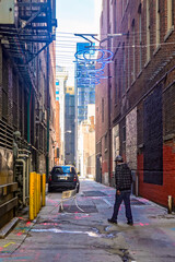 Fototapeta premium Downtown city back alley streets with skyscraper buildings and a person walking below suspended blue neon letter sign.