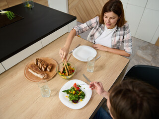 Overhead view of a beautiful young pregnant woman, attentive wife serving her husband a vegetable salad. Happy Caucasian couple enjoy togetherness over lunch