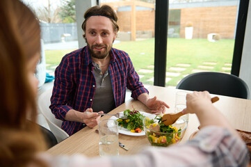 Caucasian man sits at table and has cheerful conversation with his wife, who serves him delicious healthy salad. Beautiful couple having dinner together at home against windows overlooking the garden