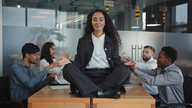 Young hispanic girl sitting on table in office in lotus position relaxing feeling calm, harmony businesswoman meditate ignore angry annoying colleagues female executive doing yoga exercises for stress