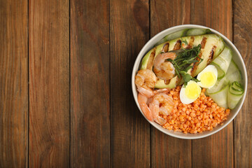Delicious lentil bowl with avocado, shrimps, egg and cucumber on wooden table, top view. Space for text