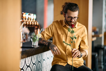 Romantic man smells red rose while waiting for his girlfriend at bar counter.