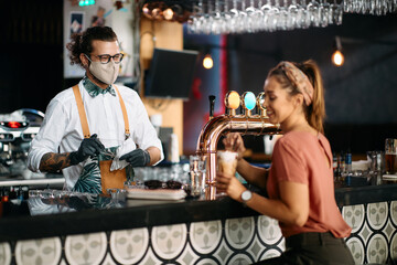 Happy bartender wears face mask while cleaning drinking glasses and talking to female guest.
