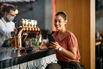 Happy woman drinks cocktail while text messaging on cell phone at bar counter.
