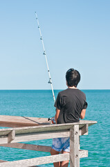 back view, medium distance of a young asian boy fishing of a woodier with wood railings, jutting into tropical Caribbean waters, on a sunny day