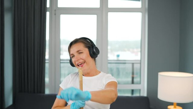 Woman in headphones cleans the house and have fun singing with a broom like a star at a concert.