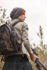 Fototapeta premium Beautiful and intense redhead sniper woman in profile with .50 caliber rifle pointed down, short hair, stocking cap, jacket, backpack looks sideways through the forest on an overcast day, low angle