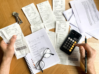 Man is making audit of household expenses using calculator and notebook. Lots of receipts and bills. Accountant, tax, vat, debt, payment, credit, finance, analysis, income, investment, cost, balance.